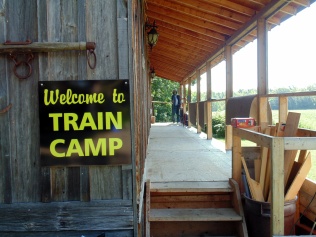 a covered porch with a figure at one end a sign on the adjoining building that says welcome to train camp